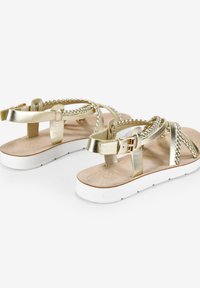 Sandalias metálicas doradas con tiras trenzadas, suela de caucho blanca y hebilla ajustable. Plantilla plana con acolchado de color marrón claro.