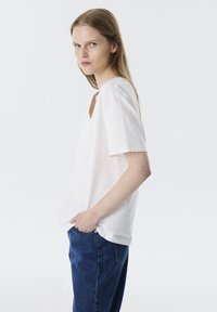 Weißes Kurzarmoberteil mit V-Ausschnitt, aus glattem Stoff gefertigt. Kombiniert mit blauen Jeans im lässigen Schnitt.