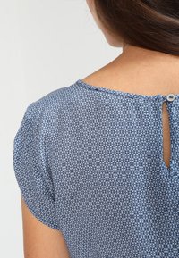 Femme portant un chemisier bleu à motifs avec de petites formes florales, fermeture par boutons au niveau de la nuque à l'arrière et manches courtes.