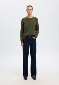 Pulover din lână de culoare verde măsliniu cu guler rotund, asortat cu blugi denim comozi de culoare bleu închis și balerini negri cu vârful ascuțit. Design simplu, material texturat.