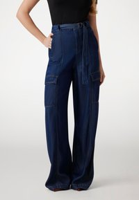 Pantaloni cargo a gamba larga in denim scuro con vita alta, passanti per cintura, tasche laterali e dettagli di cucitura visibili. Texture liscia.