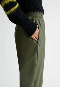 Pantalones de piel sintética verde oliva con un bolsillo lateral, que presentan una cintura elástica, combinados con un suéter de punto negro con detalles amarillos.