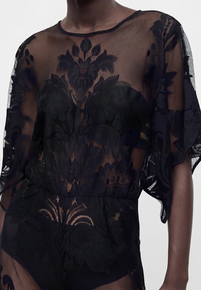 Femme portant un haut en dentelle noire transparente avec des motifs floraux et un bas assorti sur un fond uni.