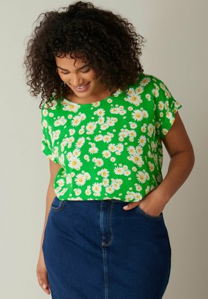 Groene bloemenblouse met witte madeliefjespatronen, korte mouwen en een relaxte pasvorm, gecombineerd met een donkere denimrok.