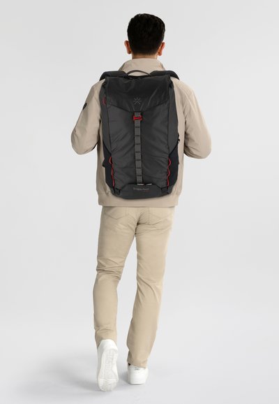 Mochila negra con acentos rojos en contraste, correas ajustables y cierre en la parte superior, combinada con un atuendo beige y zapatos blancos.