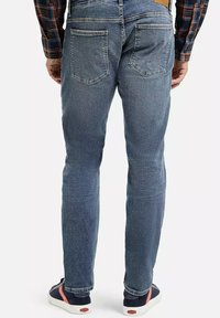 Wypłowiałe niebieskie dżinsy o slim fit, pięć kieszeni, z delikatnym efektem worn, w połączeniu z ciemnymi sneakersami z czerwonymi akcentami.