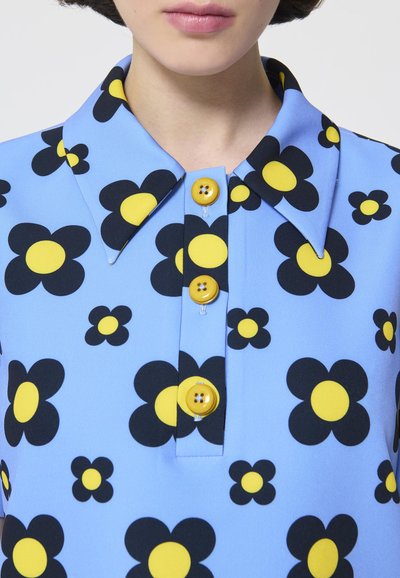 Blaue Bluse mit floralem Muster, featuring schwarze Blumen mit gelben Mittelpunkten. Vier gelbe Knöpfe auf der Vorderseite. Glatte Stoffoberfläche.