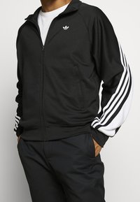 Zwarte zip-up jas met witte gestreepte mouwen, gemaakt van een zachte stof. Voorzien van een klein logo op de borst en zijzakken.