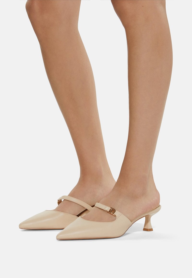 Beige lädermules med spetsig tå, med ett enstaka rem och en liten, böjd klack. Slät yta med subtil glans.