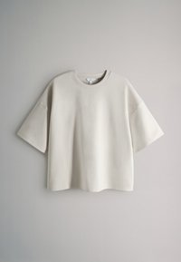 Tricou bej oversized cu mâneci scurte și decolteu rotund, atârnând pe un fundal simplu gri deschis.