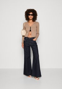Beige ribbed cardigan with button closure, black crop top, dark blue flared jeans, and white heeled sandals. Accented with a small cream bag.
