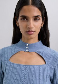 Suéter de punto azul con un cuello alto, escote con corte y tres botones decorativos. Presenta patrones texturizados y tela acanalada.
