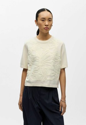 Femme aux cheveux noirs attachés en arrière, portant un pull crème à manches courtes avec un motif floral texturé et un pantalon noir ample à rayures fines, debout en position neutre.