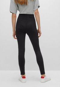 Leggings noirs taille haute en tissu lisse, avec une taille fermée et des jambes fuselées. Portés avec des baskets blanches.