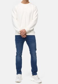 Felpa color crema con collo rotondo e polsini a costine, abbinata a jeans blu e scarpe da ginnastica bianche. La felpa presenta una piccola toppa con logo.