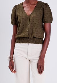 Blouse vert olive à manches courtes avec un col en V, un motif tricoté texturé, une fermeture à boutons et un ourlet smocké. Associée à un pantalon de couleur crème.