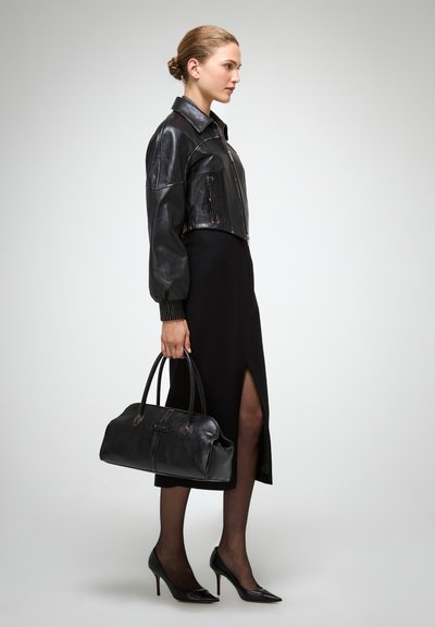 Chaqueta corta de cuero negro con cierre de cremallera, combinada con un vestido midi negro con una abertura lateral. Los accesorios incluyen un gran bolso de mano negro.