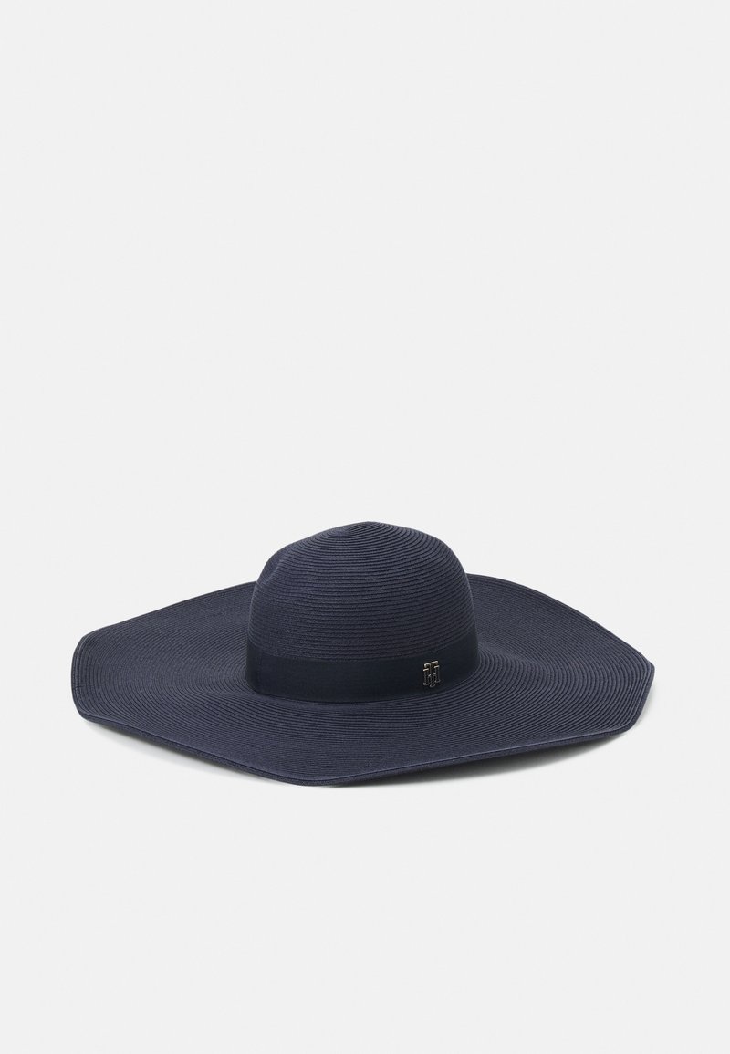 Chapeau de soleil bleu marine foncé à larges bords, orné d’un ruban noir et d’un petit détail métallique de logo sur le ruban, présenté sur un fond uni clair.