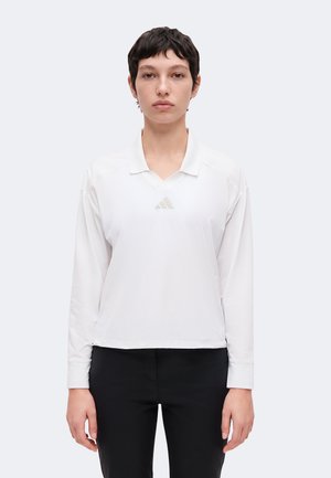 Mladá osoba s krátkymi tmavými vlasmi má na sebe biele polo s dlhým rukávom s jemným logom a čierne nohavice, stojí pred jednofarebným pozadím.