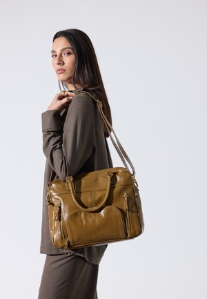 Woman in brown outfit carrying large tan leather shoulder bag with multiple zippered pockets against plain white background.