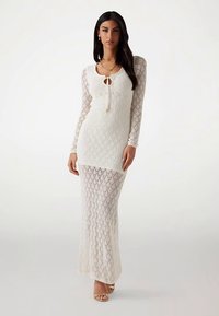 Weißes gestricktes Maxikleid mit langen Ärmeln, Schlitz am Ausschnitt mit Bindeband, durchgehendem Rautenmuster und transparentem Stoff im unteren Bereich.