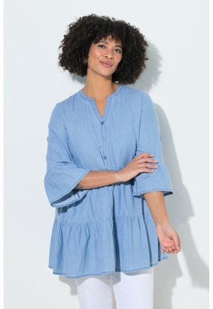 Kvinde med krøllet hår iført en løs, lyseblå knapbluse med trekvartærmer og hvide bukser, stående mod en ensfarvet baggrund.