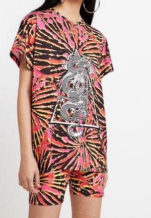 Nainen pukeutuneena eläväväriseen tie-dye -paitaan ja shortseihin, joissa paidan etupuolella oleva valkoinen kolmio sisältää mustavalkoisen lohikäärmekuvion.