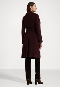 Cappotto aderente di un intenso bordeaux, con ampio revers, vita con cintura e spacco posteriore. Abbinato a pantaloni in velluto marrone e stivali con tacco.