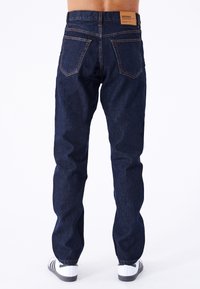Jeans de mezclilla azul oscuro con un corte de pierna recta, bolsillos traseros visibles, costuras naranjas y una etiqueta marrón en la cintura.