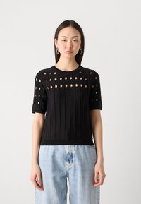 Black knit top featuring horizontal ribbing and circular cut-outs, short sleeves, and a fitted waistband, paired with light denim jeans.