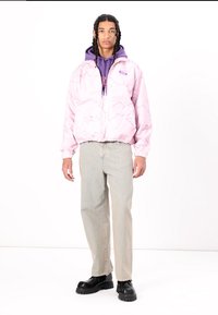 Light pink bomber jacket with abstract patterns, zip closure, and elastic cuffs worn over a purple hoodie. Paired with light jeans and black shoes.