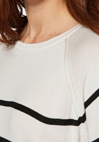 Maglietta a maniche lunghe color crema con strisce nere. Presenta un colletto rotondo a coste e una morbida texture a maglia. Cuciture a vista sulle spalle.
