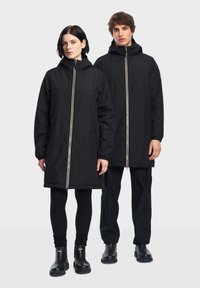 Giubbotti neri impermeabili con cappucci, dotati di zip frontali e tasche laterali, indossati da due figure in pantaloni e stivali neri abbinati.