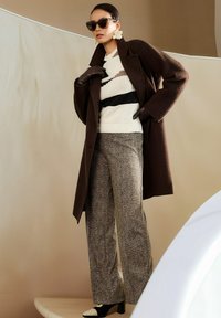 Frau trägt einen dunkelbraunen Mantel, gemusterte weite Hose, gestreiften Pullover, Lederhandschuhe, große Blumenohrringe, Sonnenbrille und schwarz-weiße High Heels.