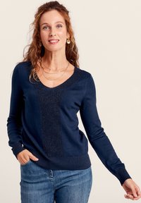Dunkelblauer Pullover mit V-Ausschnitt, strukturiertem Vorderpanel und gerippten Bündchen sowie Saum. Getragen mit hellblauen Jeans und langen Ärmeln.
