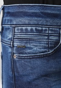 Jeans de mezclilla en azul oscuro con una textura desgastada. Presenta un bolsillo trasero con un acento de logotipo metálico y detalles de costura.