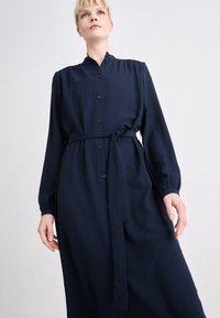 Navy blue, long-sleeved dress with a collar, front buttons, and a waist tie. The fabric appears textured with a soft drape and relaxed fit.