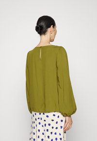 Blouse vert olive avec un col rond, des manches bouffantes et un détail en trou de serrure dans le dos, associée à une jupe crème avec des taches bleues.