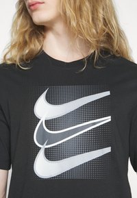 Černé bavlněné tričko s grafickým potiskem tří překrývajících se Nike swooshů v různých odstínech šedé na tečkovaném pozadí.