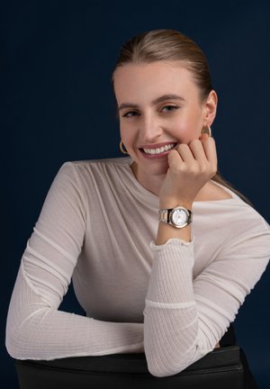 Reloj en tonos plateado y dorado con un dial blanco y pulsera de metal, llevado en la muñeca de una mujer. Parte superior de manga larga ligeramente texturizada en color pálido.