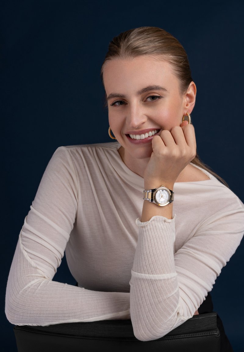 Orologio in tonalità argento e oro con quadrante bianco e bracciale in metallo, indossato al polso di una donna. Top a maniche lunghe leggermente strutturato in colore chiaro.