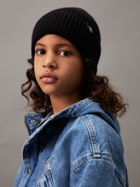 Gorro negro de canalé con un pequeño logo, combinado con una chaqueta de mezclilla azul con cremallera y detalles bordados. Textura suave, corte casual.