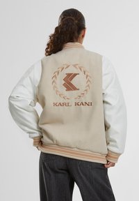 Bēžs bomberjaka ar baltām piedurknēm, ribotām aprocēm un apkakli. To raksturo izšūts logo un lauvas dizains uz aizmugures.