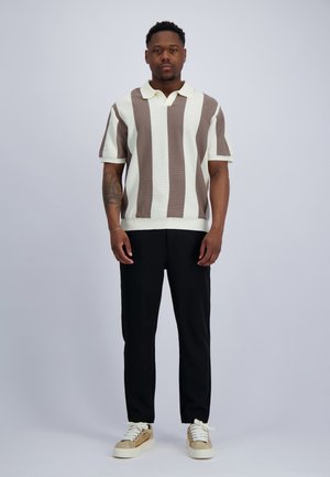 Man staat, draagt een kort mouw gebreide polo met verticale beige en witte strepen, zwarte broek en beige sneakers met witte veters.