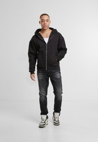 Sudadera negra con cremallera y bolsillos frontales, llevada sobre una camiseta sin mangas blanca, combinada con pantalones oscuros y zapatillas altas con un diseño a rayas.