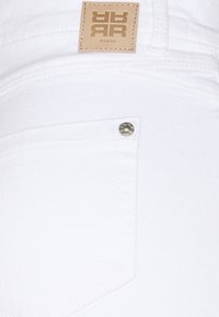 Bílé denimové džíny s malou přední kapsou a viditelnou značkovou štítkou v hnědé kožené úpravě s embosovaným logem.