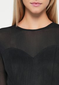 Žena v černém topu s průhledným výstřihem a neprůhledným korzetem ve tvaru srdce, rty mírně pootevřené.