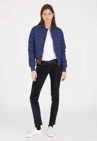 Giacca puffer blu navy con zip frontale e polsini a costine, abbinata a pantaloni neri in tessuto testurizzato e sneakers bianche. Design semplice e casual.