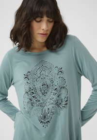 Langermige teal shirt met een zwart versierde mandala-ontwerp op de voorkant. Zachte stof met een gladde textuur en een comfortabele pasvorm.