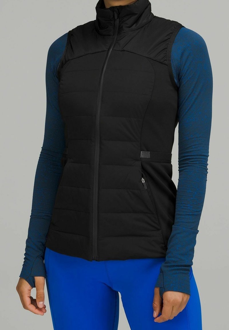 Personne portant un gilet noir matelassé sans manches sur une chemise bleue cintrée à manches longues avec des passages pour les pouces et un pantalon bleu vif.